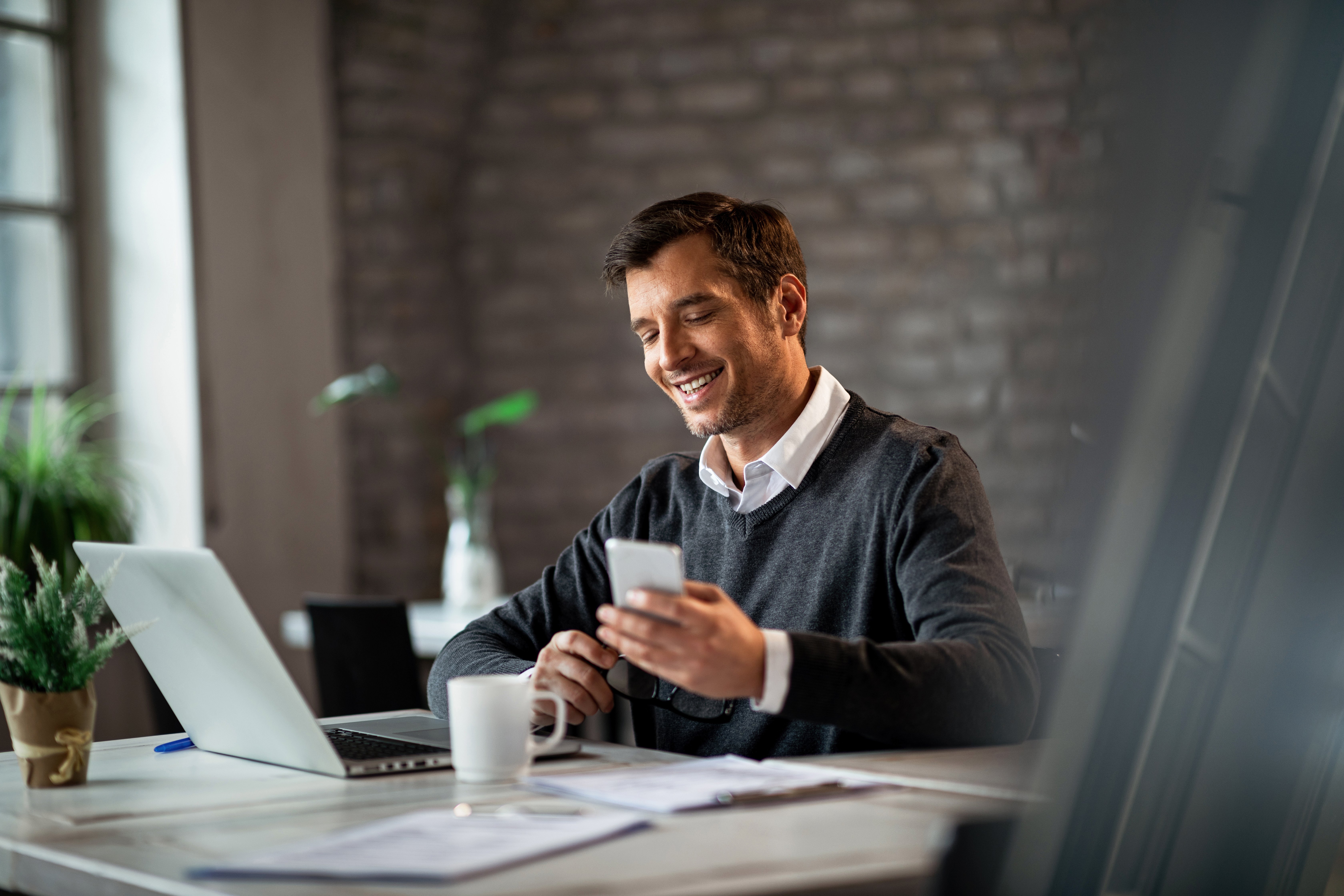 happy-businessman-texting-mobile-phone-while-working-laptop-office